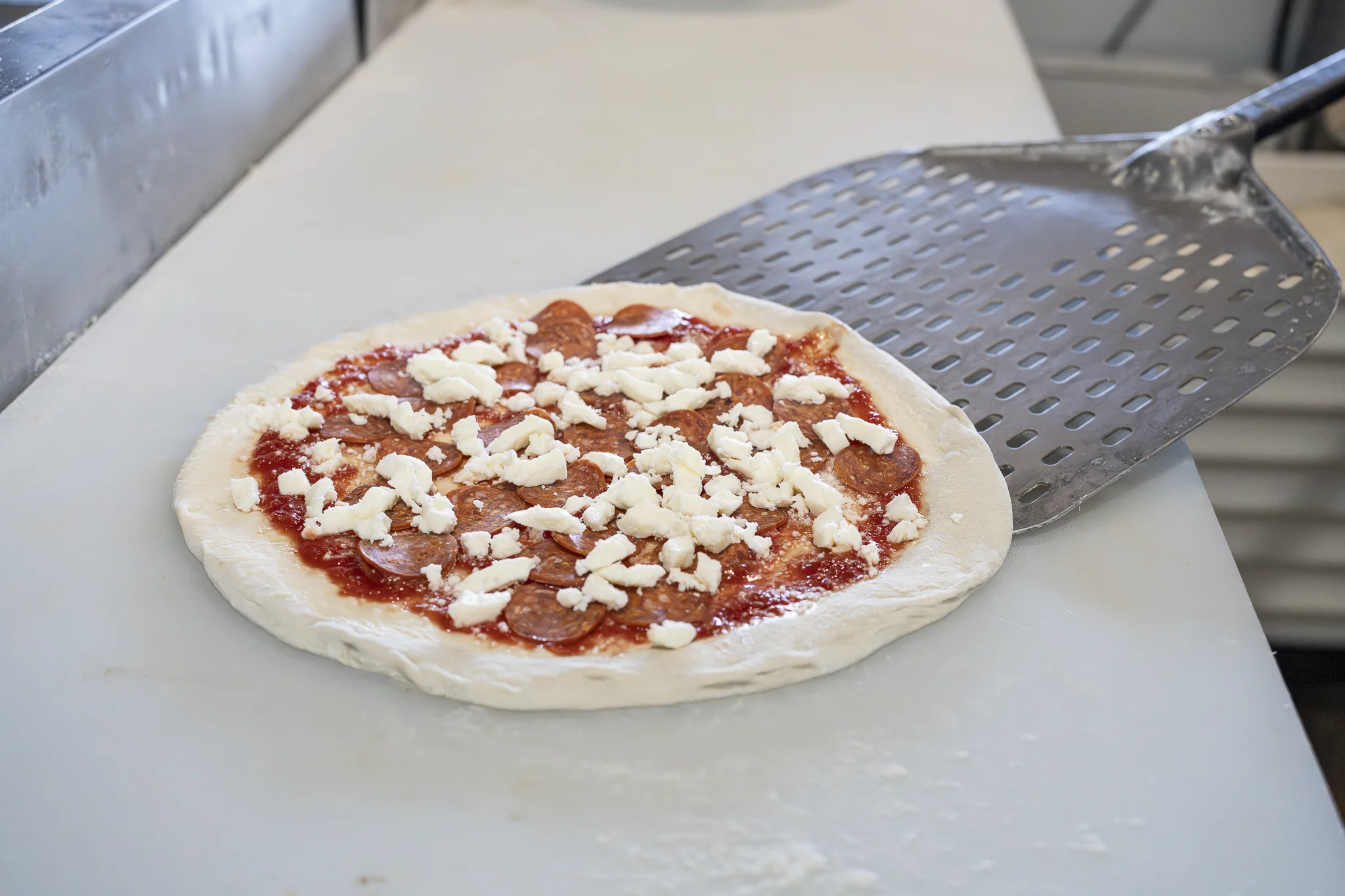 Raw pizza topped with tomato sauce, sausage, and fresh mozzarella next to a pizza peel